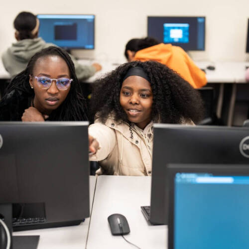 Two females look at the monitor of a computer with one pointing to it. Other computers are visible within the room that looks like a computer lab.